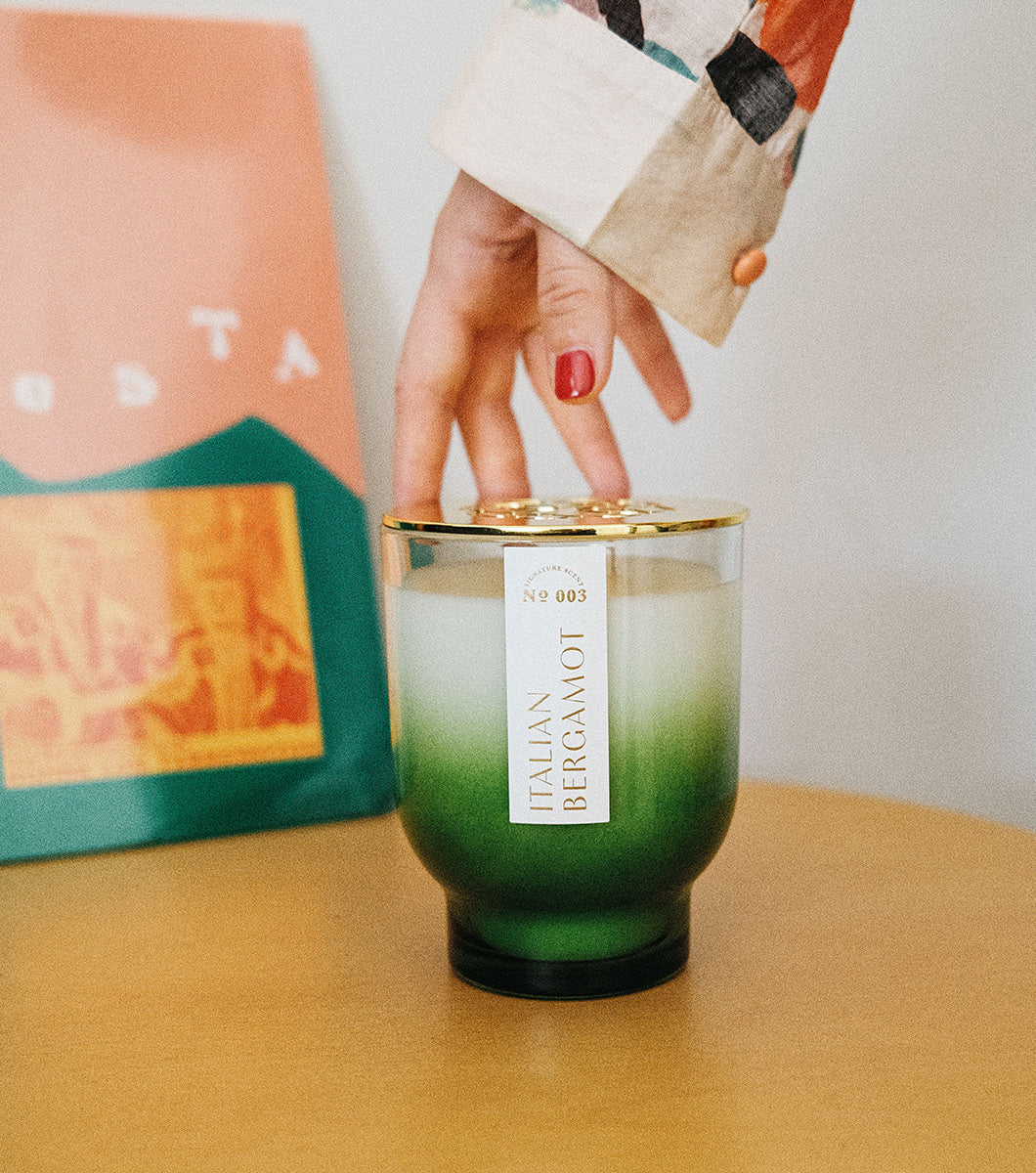 large green italian bergamot scented candle pictured with modern living space and a hand adjusting the gold lid