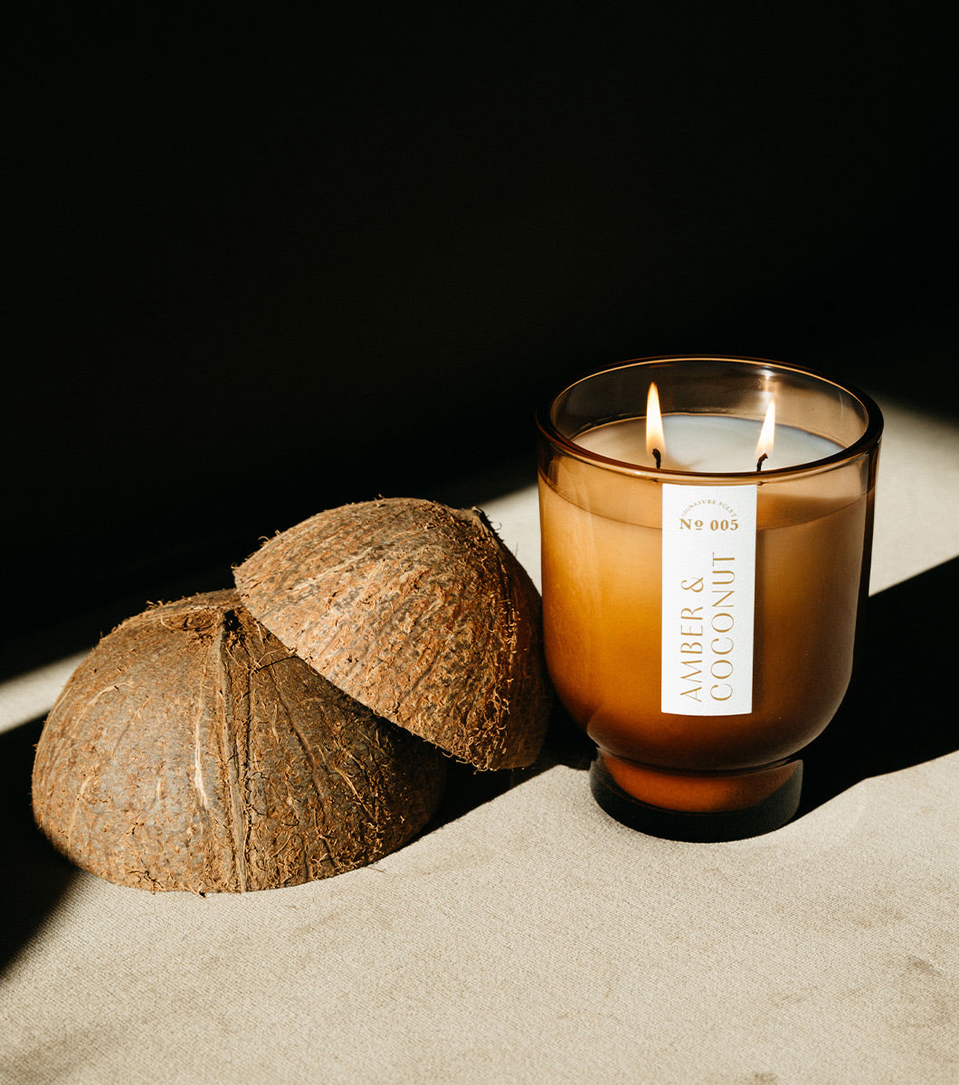 large brown amber and coconut scented candle wit hboth wicks lit pictured next to two coconut husks with a darkened shawdow background