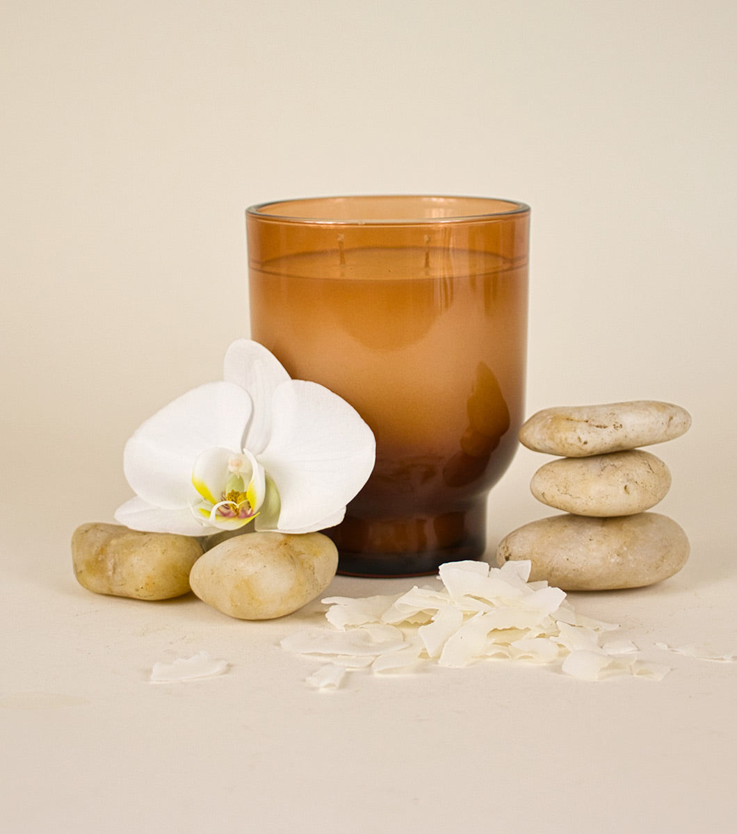 large brown amber and cocout scented candle pictured with rounded stones, white orchid and shaved coconut