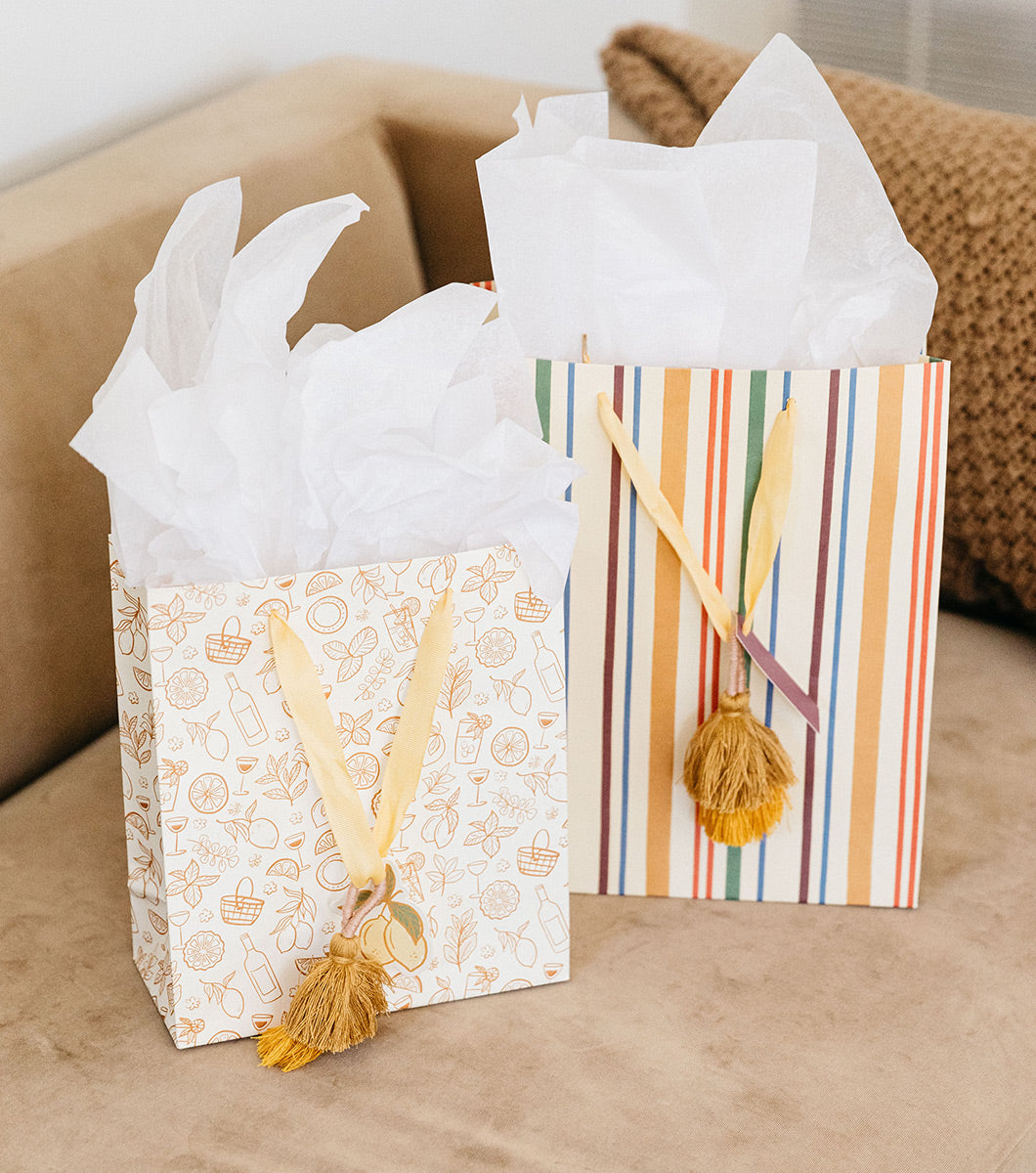 large and medium gift bags pictured on a modern brown couch. large gift bag features striped design and medium gift bag features line drawing designs of a picnic aesthetic