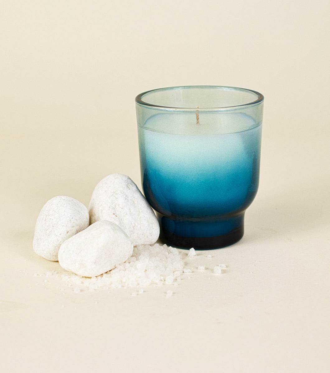 small blue serene sea salt scented candle pictured next to white stones and coarse rock salt