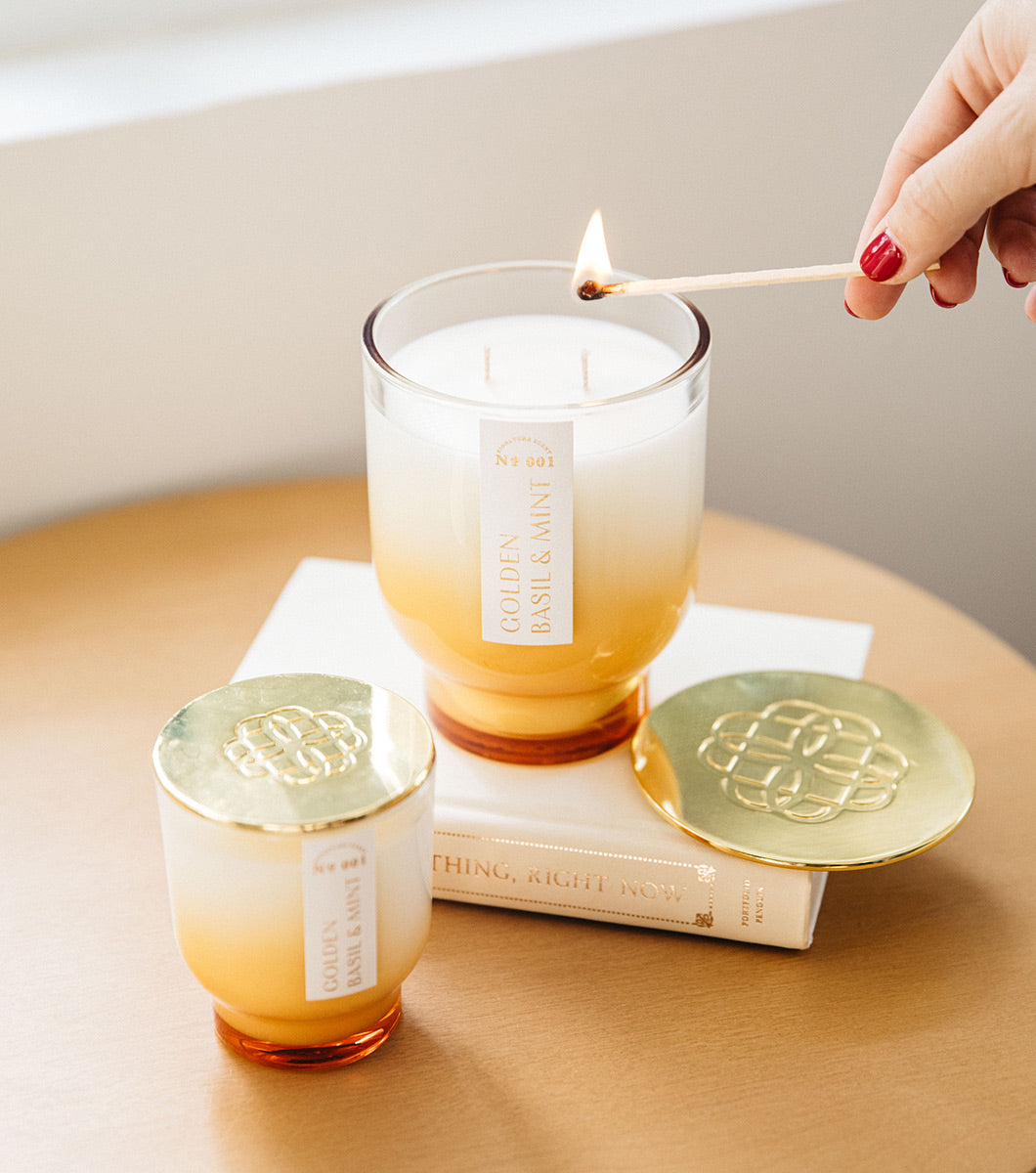 large and small yellow golden basil and mint scented candles pictured with lids and a hand is holding a match to light the larger of the candles