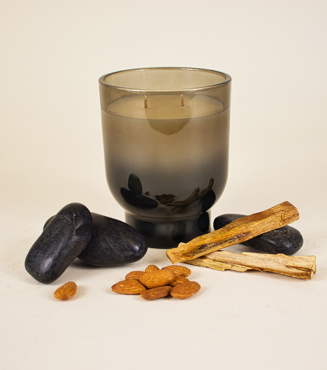 large gray and black velvet sandalwood scented candle pictured with rounded black rocks, sandalwood sticks and almonds