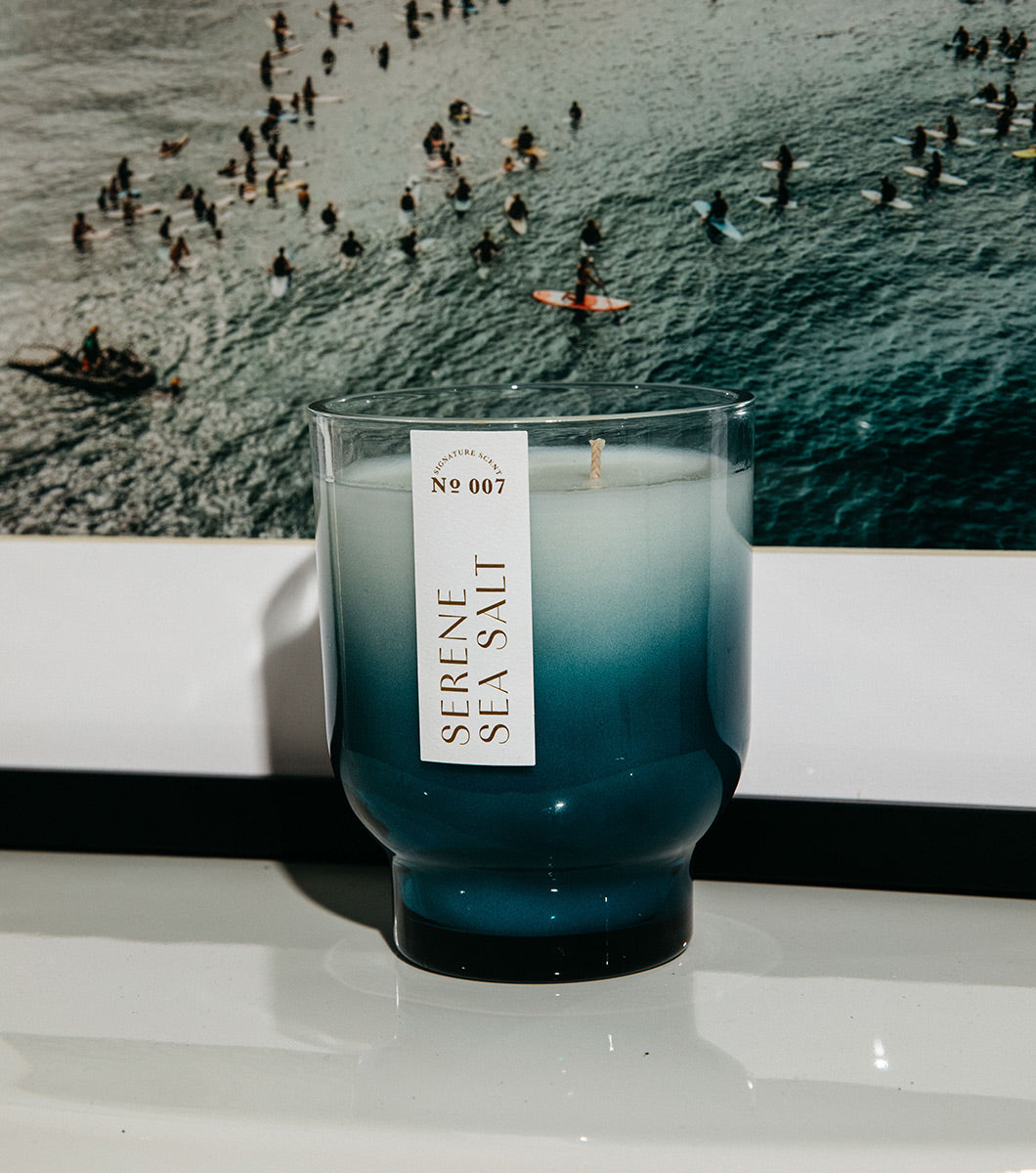 large blue serene sea salt scented candle pictured in a modern home with a photo of surfers in the background