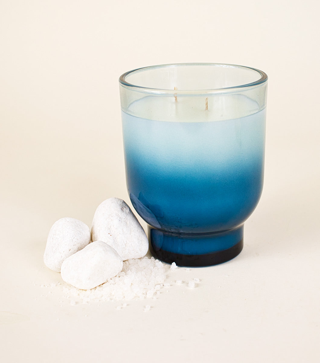 large blue serene sea salt scented candle pictured with white rocks and a small pile of coarse sea salt