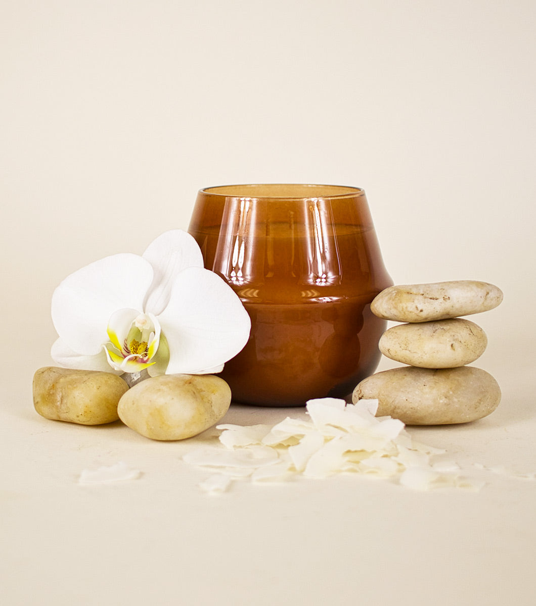 large brown amber and coconut scented  candle pictured with rounded stones, a white orchid and chaved coconut