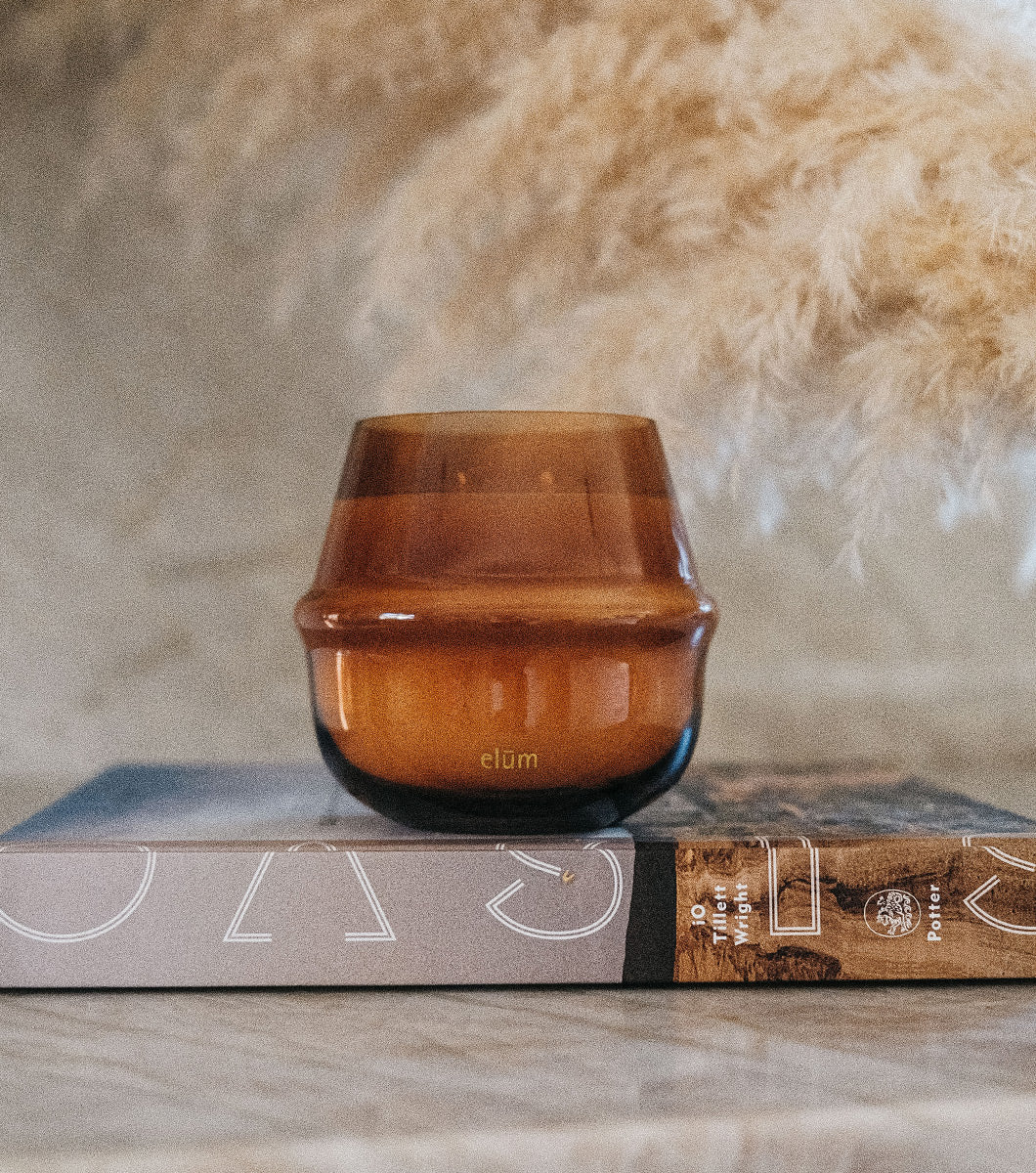 large brown amber and coconut scented candle pictured in a modern home on top of a coffee table art book and fluffy reeds in the background