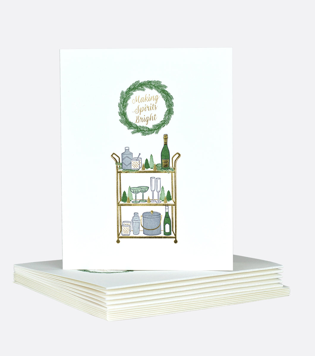 Holiday greeting card with gold bar cart with christmas decorations and alcoholic spirits and glasses, wreath above says "making spirits bright" in gold foil