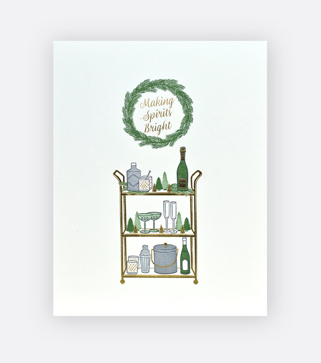 Holiday greeting card with gold bar cart with christmas decorations and alcoholic spirits and glasses, wreath above says "making spirits bright" in gold foil