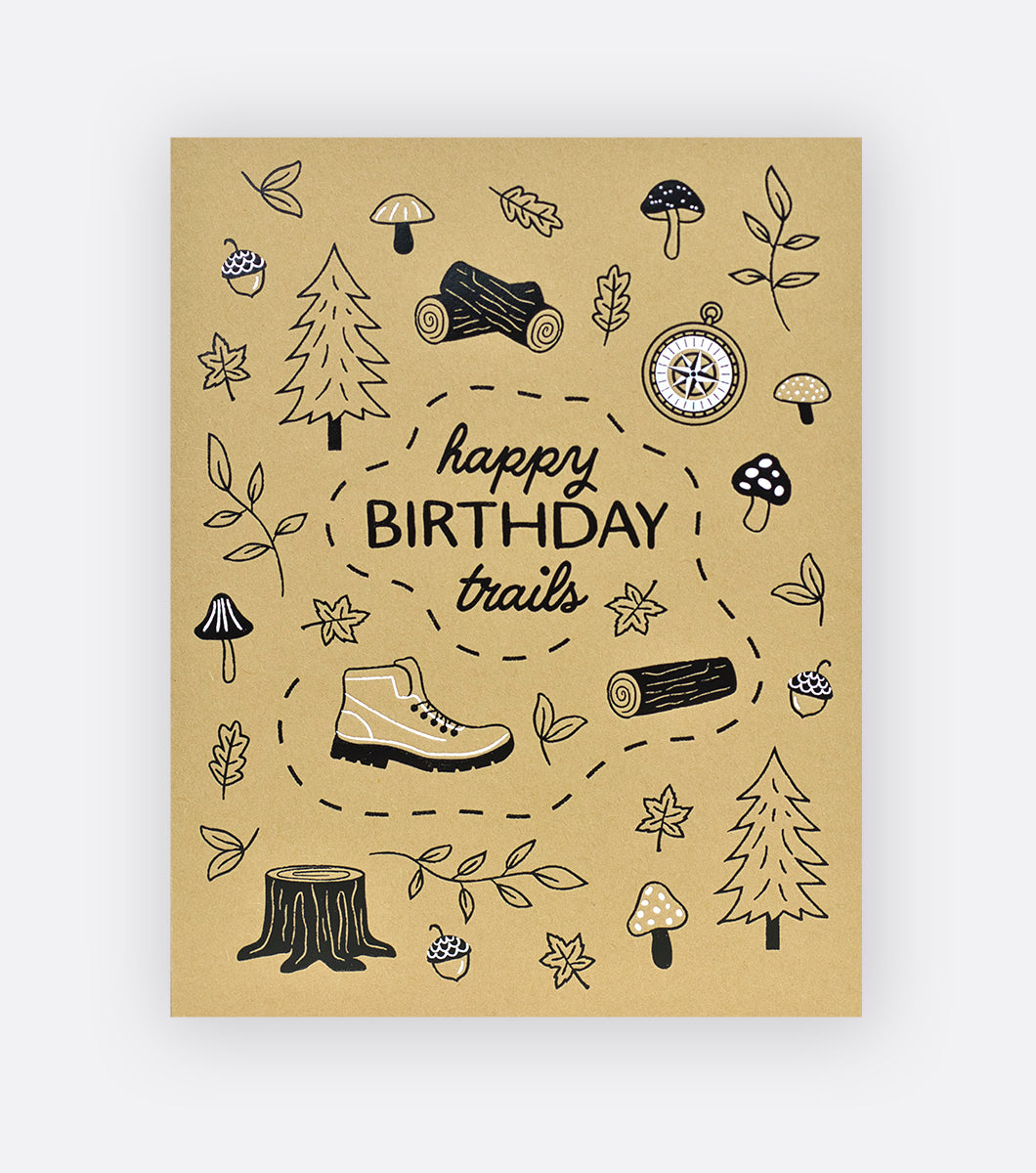 birthday greeting card on kraft paper with a white and black foil design that says "Happy Birthday Trails" with an outdoor motif of leaves and trees, hiking boots, compass mushrooms, and acorns